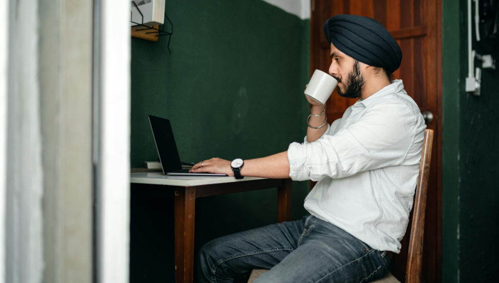 Man in turban working on laptop from home, sipping tea for productivity.